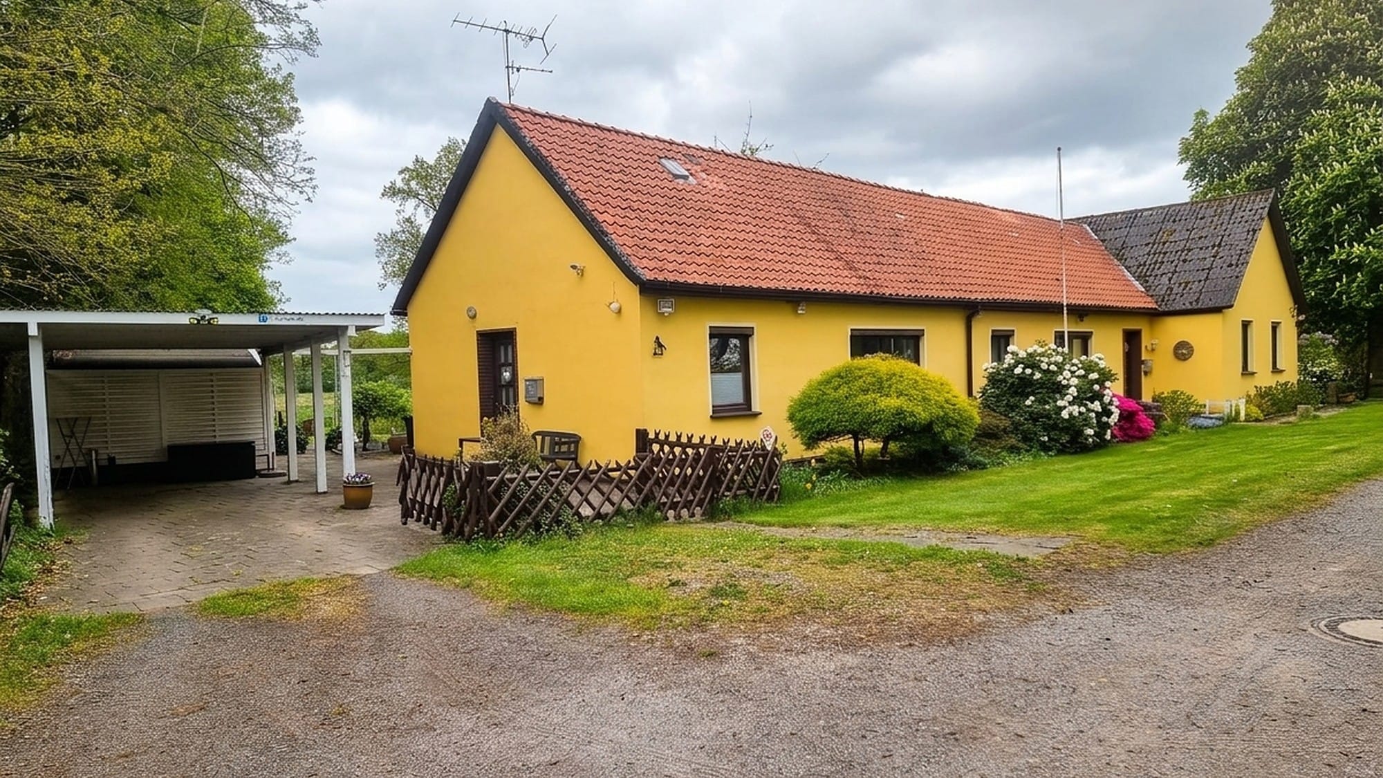 Gelbes Haus mit rotem Ziegeldach, links der Carport, davor Rasen und ein Holzzaun