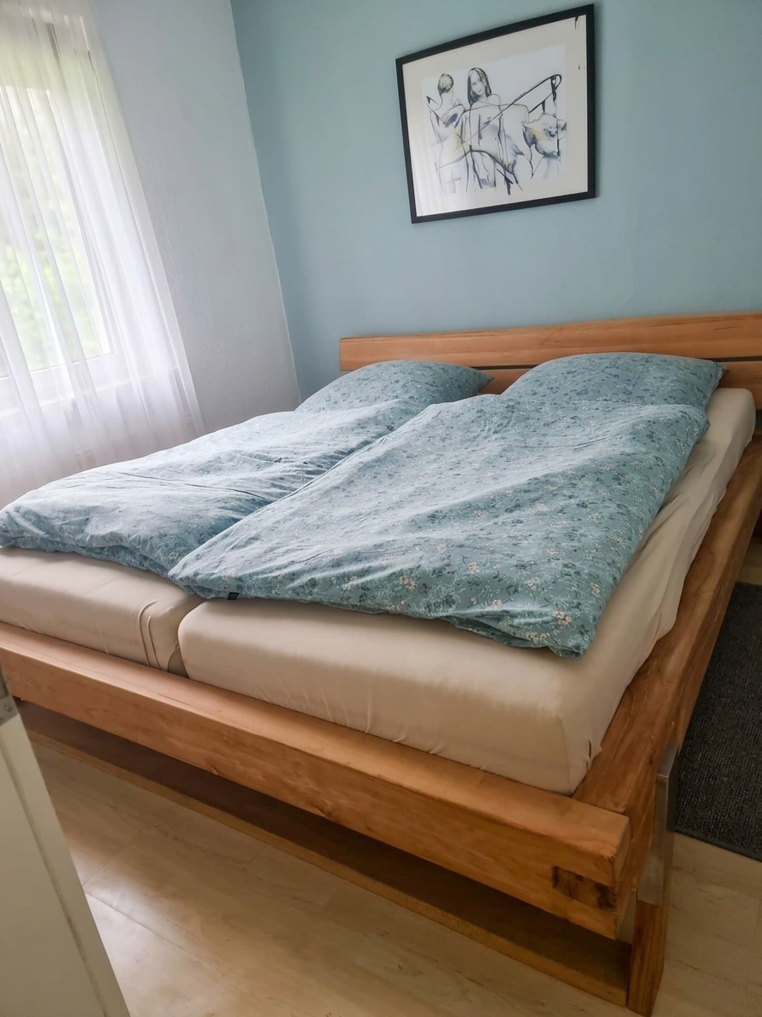 Schlafzimmer mit breitem Holzbett, geblümter Bettwäsche in Blaugrün, Bild über dem Bett und hellem Vorhang