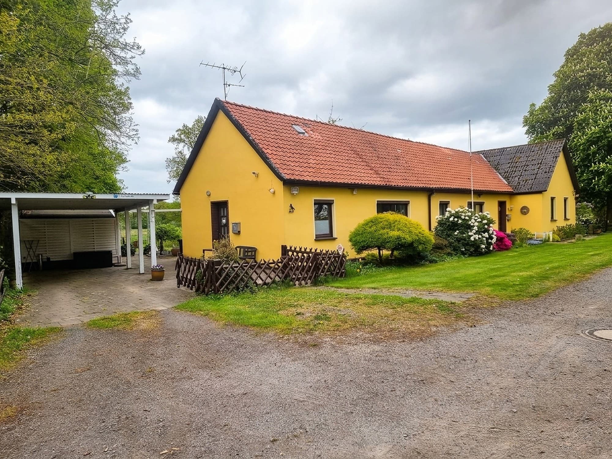 Gelbes Haus mit rotem Ziegeldach, links ein Carport, davor Rasen, Büsche und ein Holzzaun
