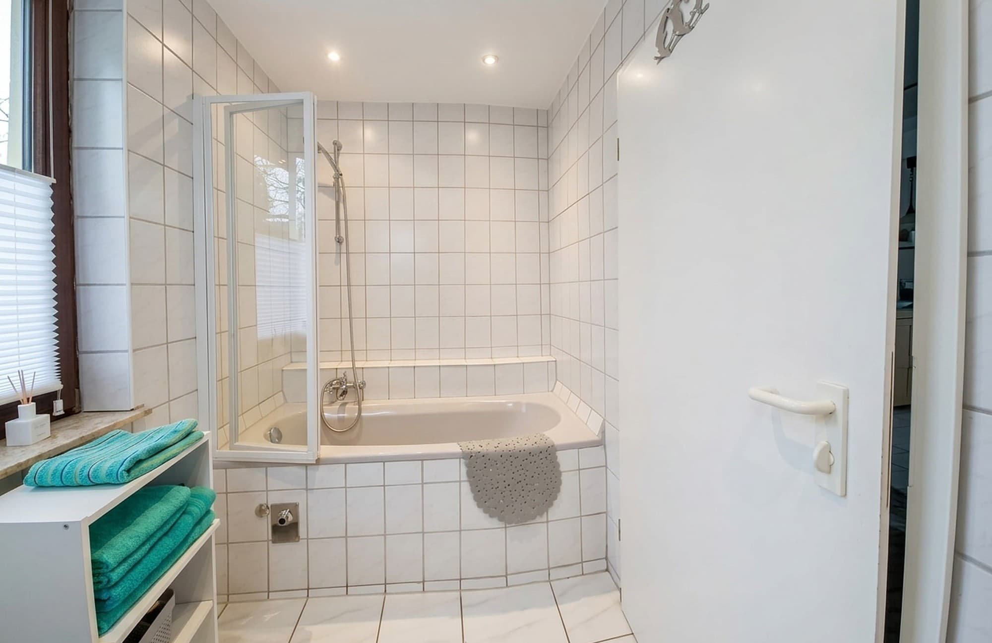 Gefliestes Bad mit Badewanne, Duschabtrennung aus Glas, Handbrause und Regal mit türkisen Handtüchern
